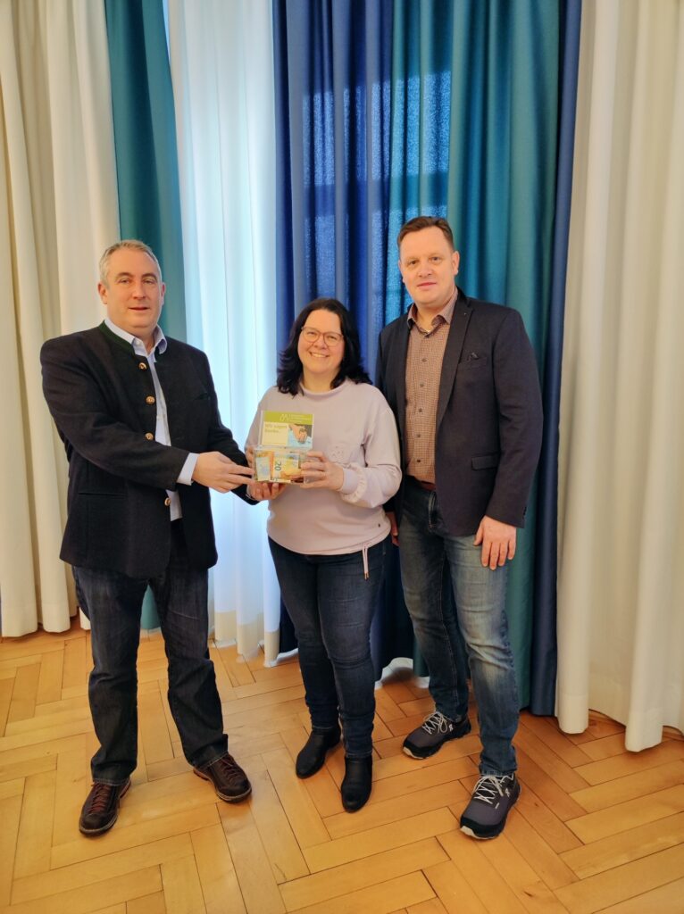 Frau des Kinderhospiz Polling zeigt Buch, zwei Männer lächeln.