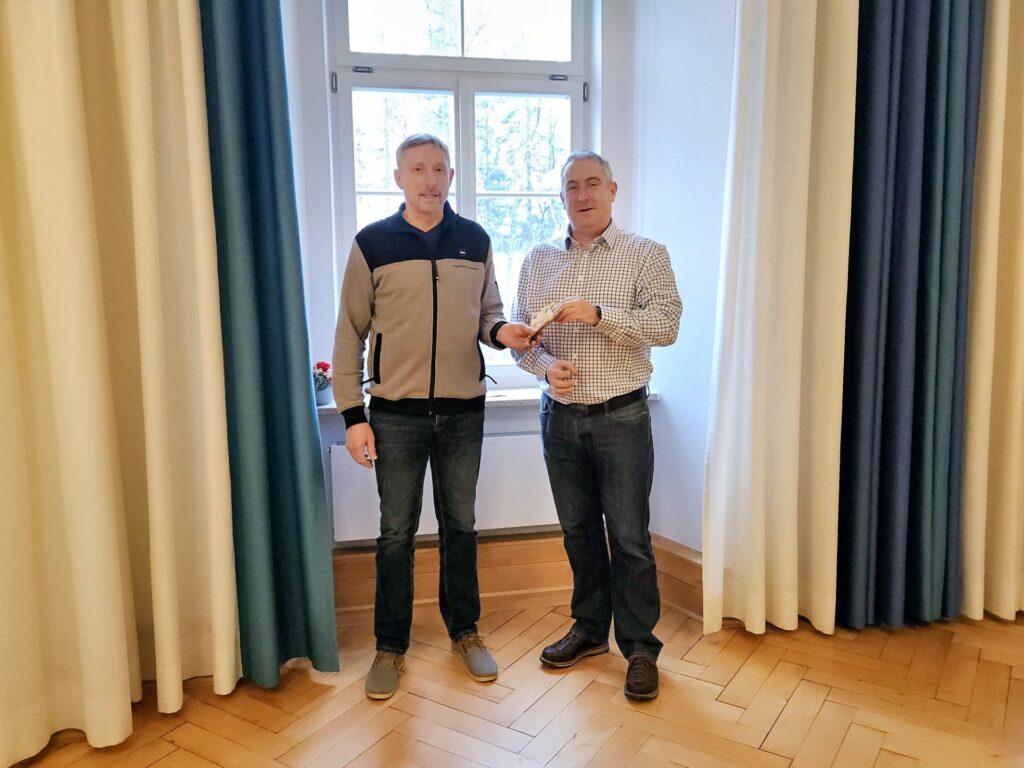 Gemütlicher Aufenthaltsraum, zwei Männer überreichen sich lächelnd einen Gegenstand.