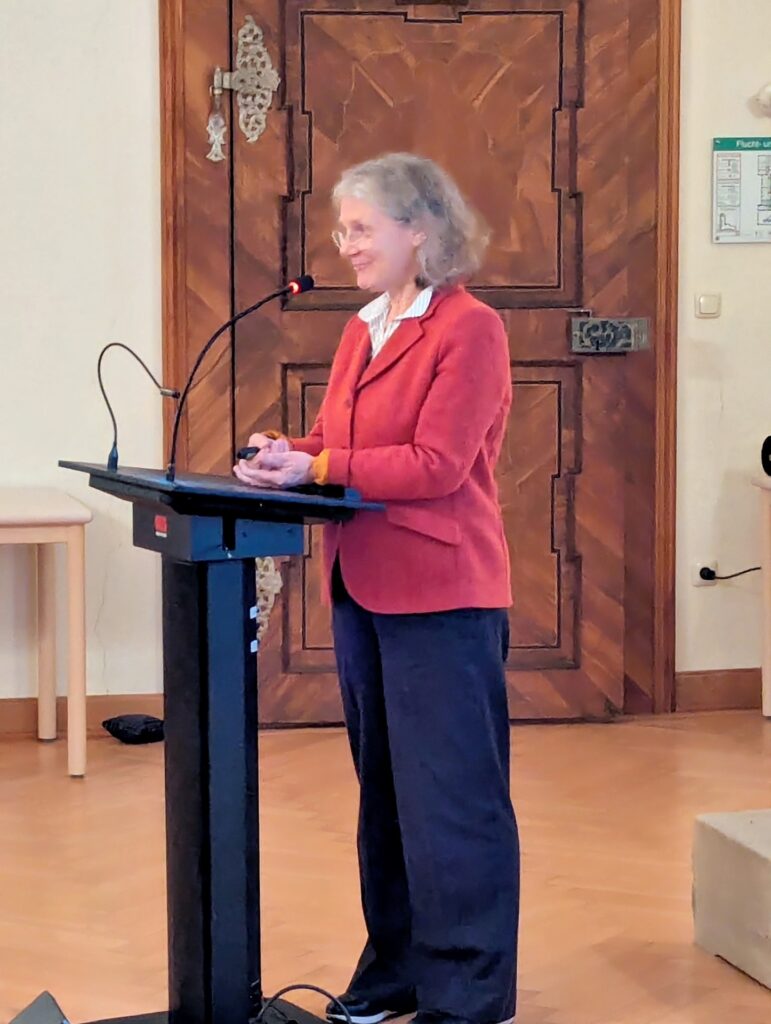 Rednerin mit grauen Haaren spricht am Podium, holzgetäfelter Saal.