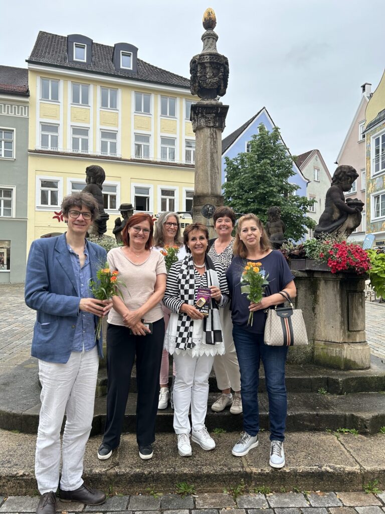 Hospiz Polling-Team mit Blumensträußen vor historischem Brunnen, Stadtplatz.