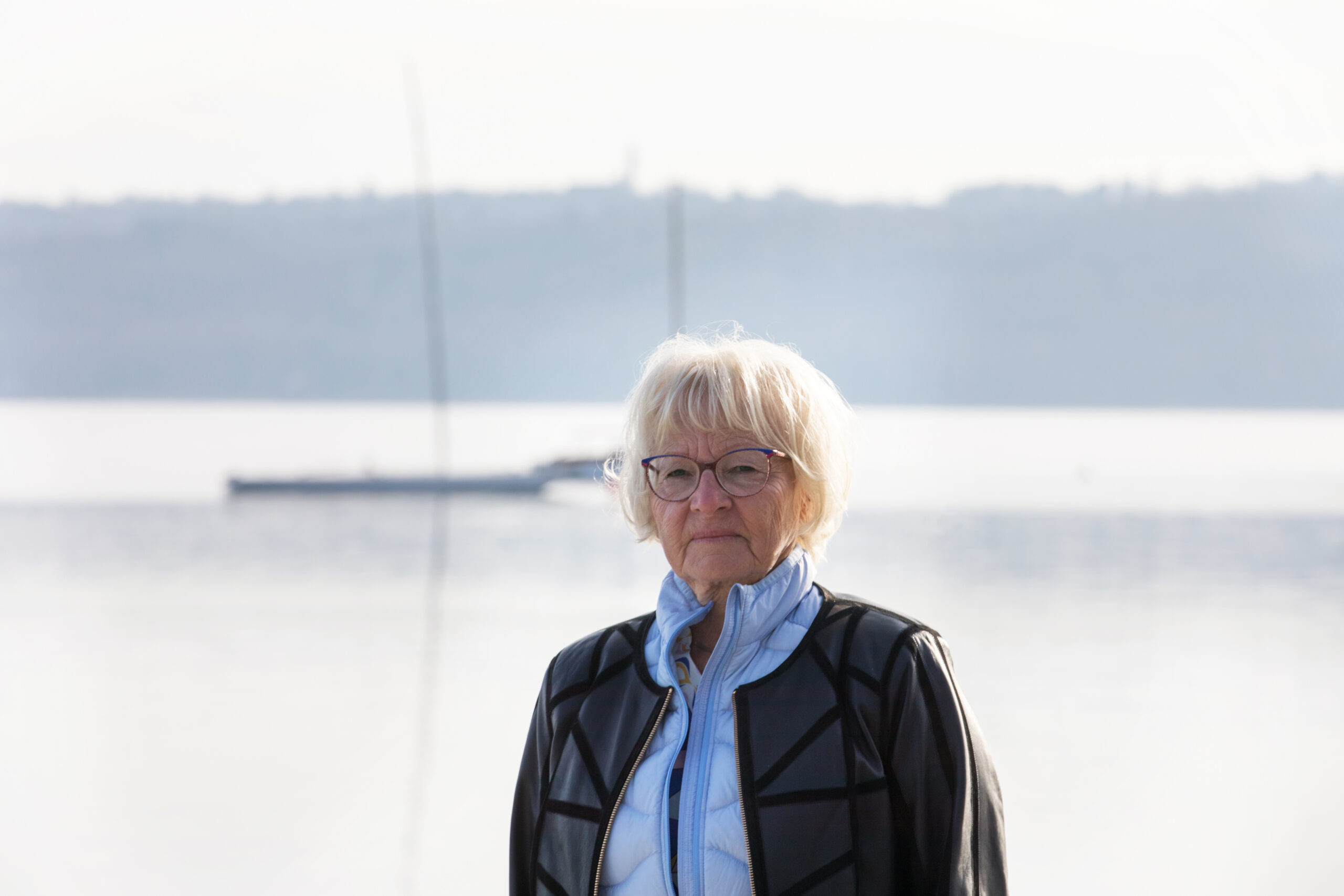 Maria mit Brille und blauem Schal am ruhigen Seeufer.